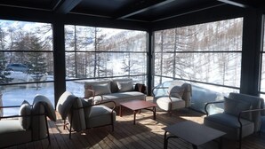 Een moderne, goed verlichte kamer met grote ramen en uitzicht op een besneeuwde berg. De kamer is ingericht met een bank, fauteuils en een salontafel, en is ontworpen voor ontspanning.