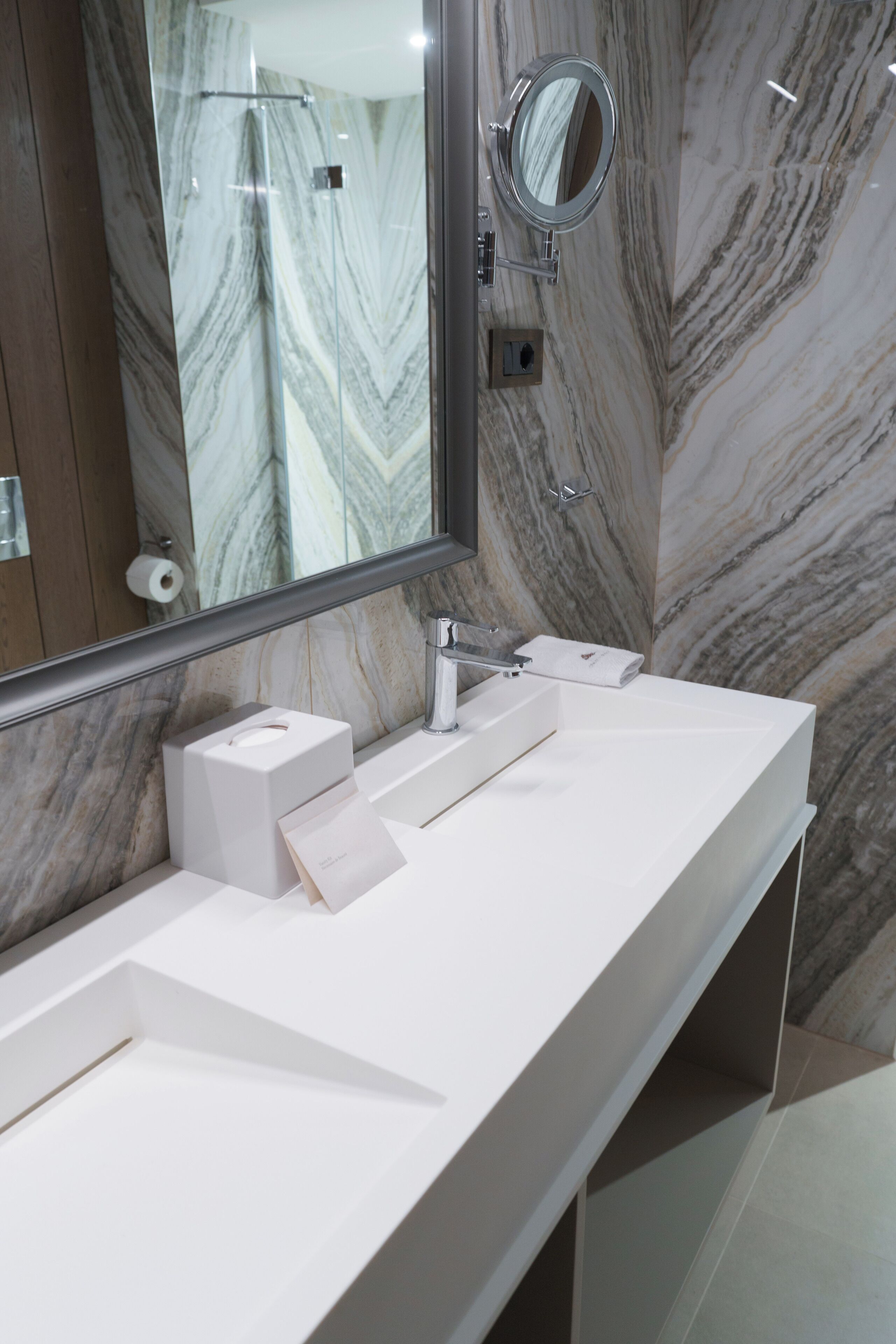 A bathroom with a marble wall, a large mirror, a white sink, and a towel dispenser.