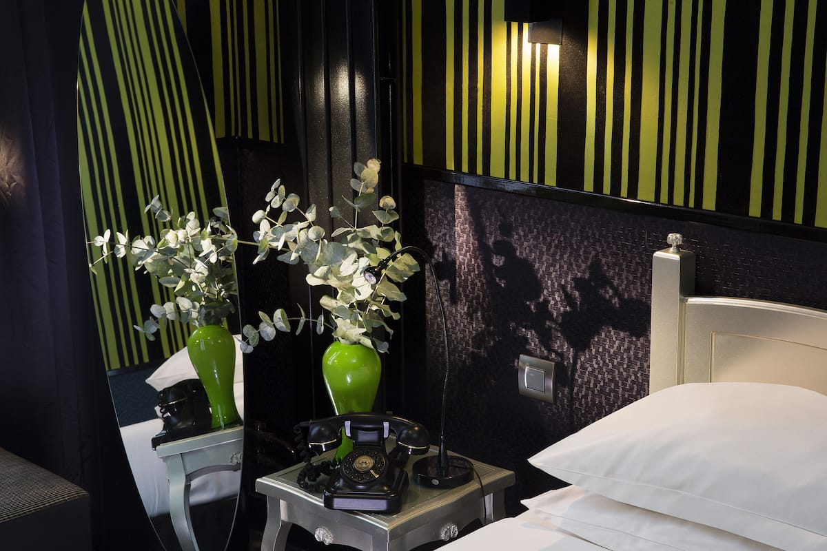 A hotel room with a bed, a bedside table with a telephone, a green vase with white flowers, and a mirror reflecting the room.