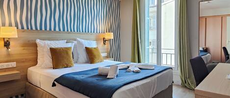 A hotel room with a large bed, two bedside lamps, a desk, and a view of the city through the window.