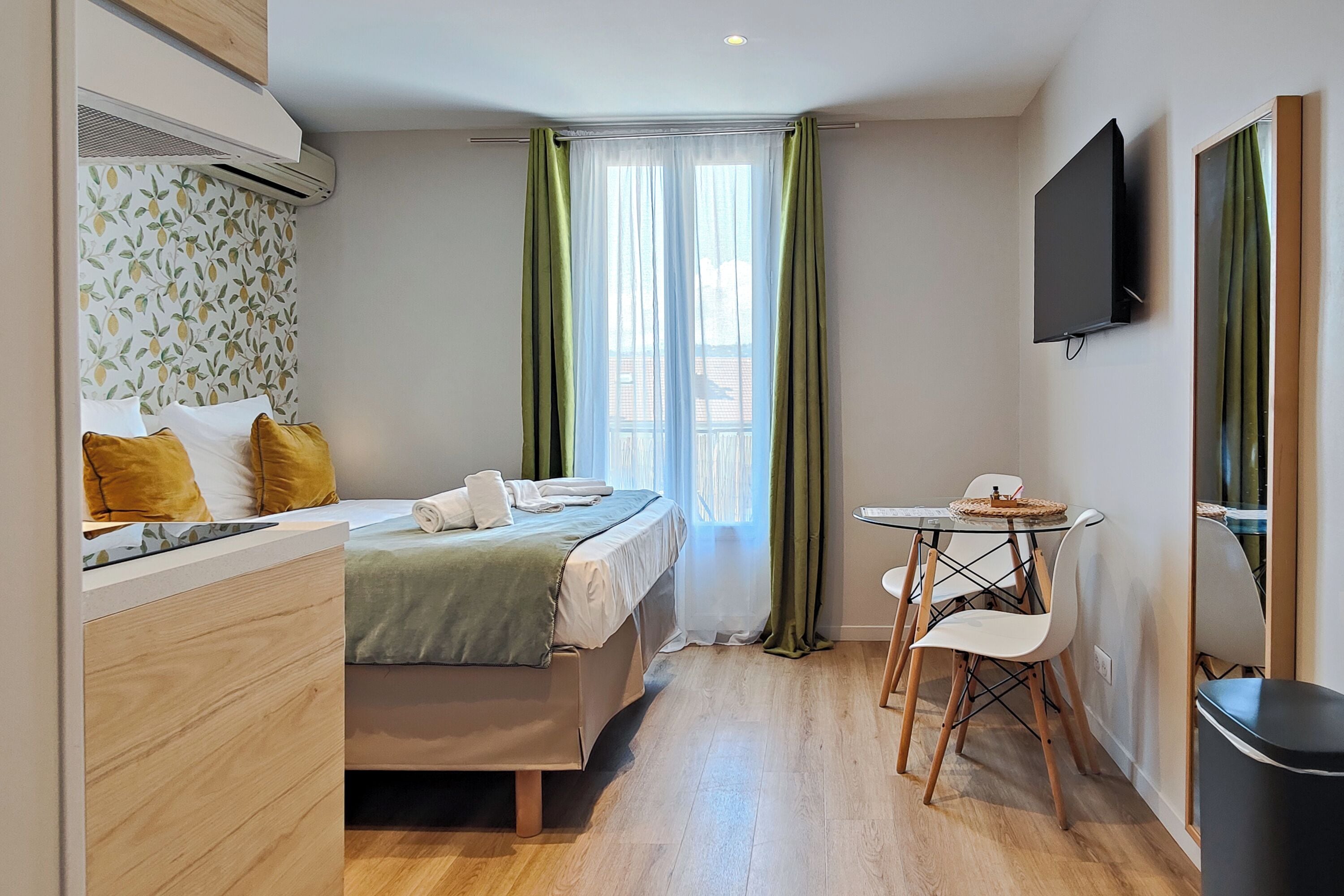Habitación de hotel pequeña con cama, una mesita con sillas, televisor montado en la pared y ventana con cortinas verdes.