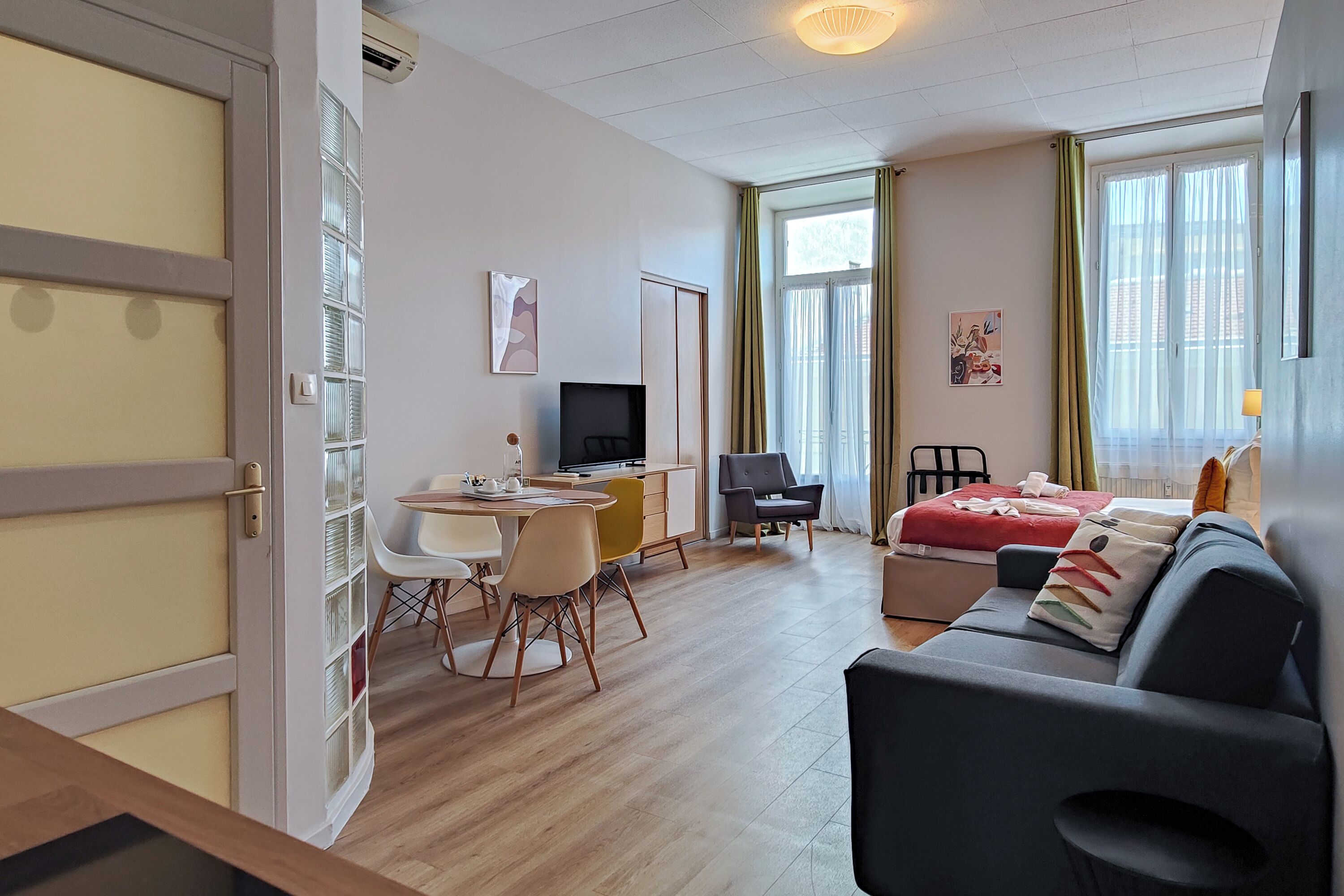 A modern hotel room with a dining area, a sofa, a TV, and large windows with curtains.