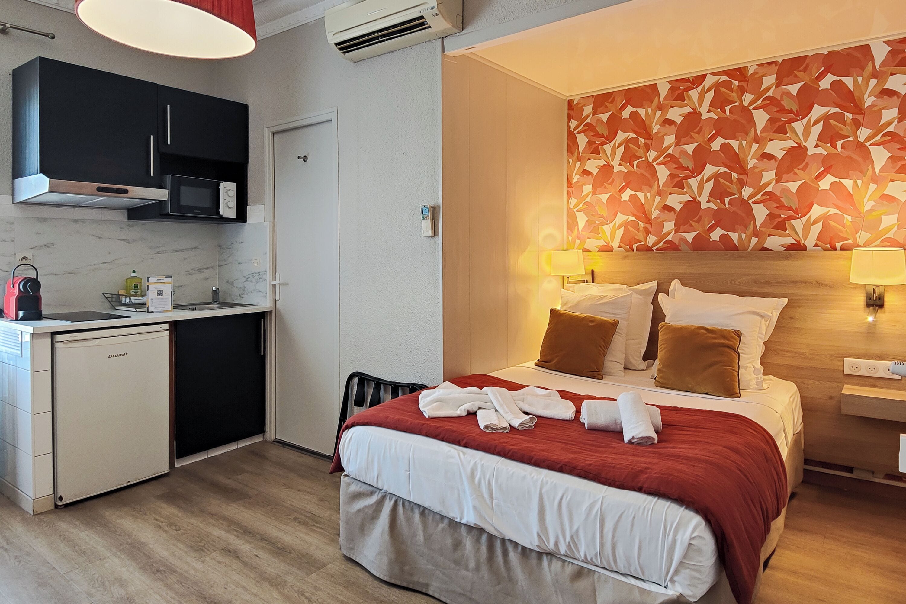 Une chambre d’hôtel équipée d’un lit, de lampes de chevet, d’une couverture rouge, d’un four à micro-ondes, d’un petit réfrigérateur et d’un coin cuisine avec des placards et un évier.