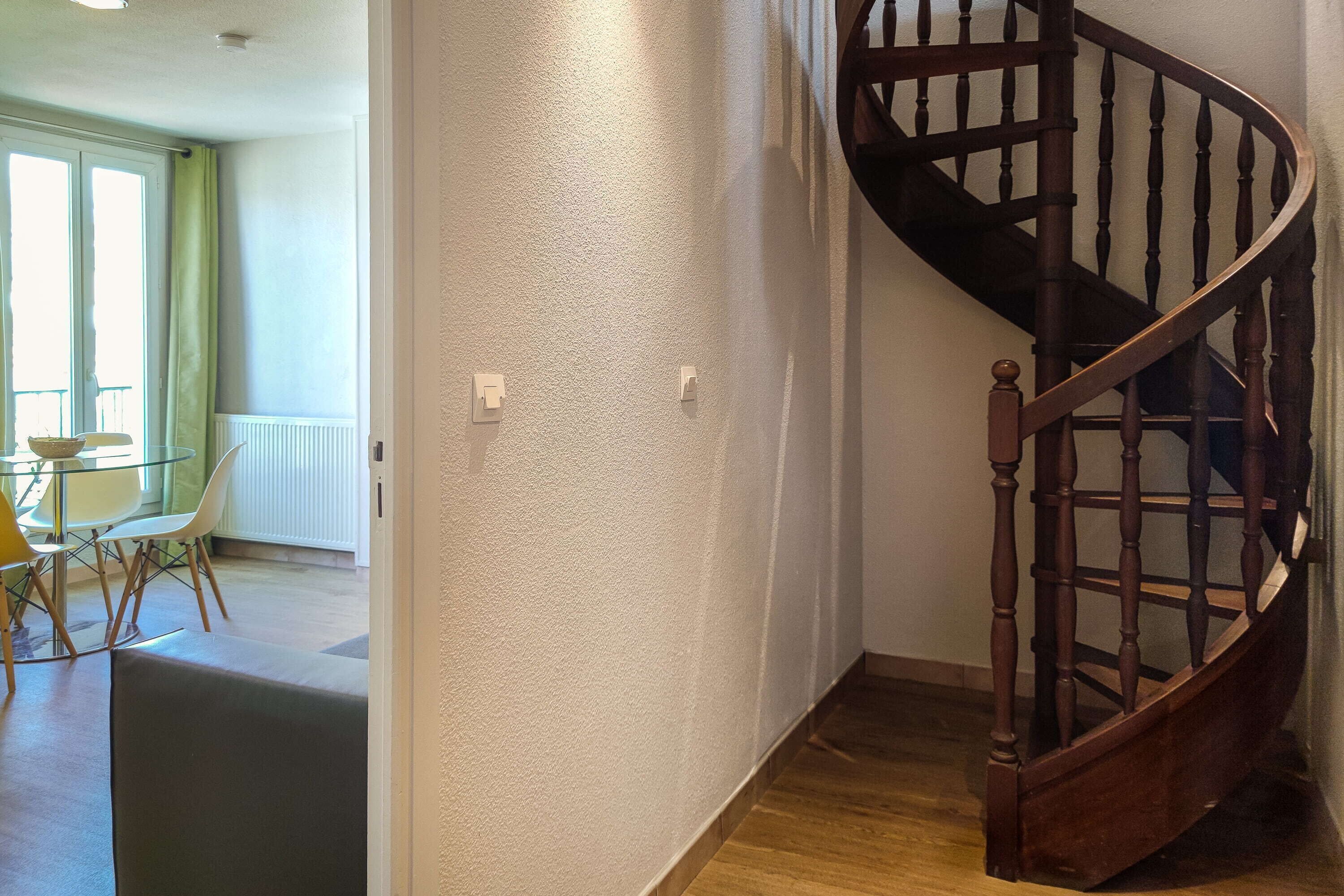 A wooden staircase with a handrail, a dining area with a table and chairs, and a view of a window with curtains.