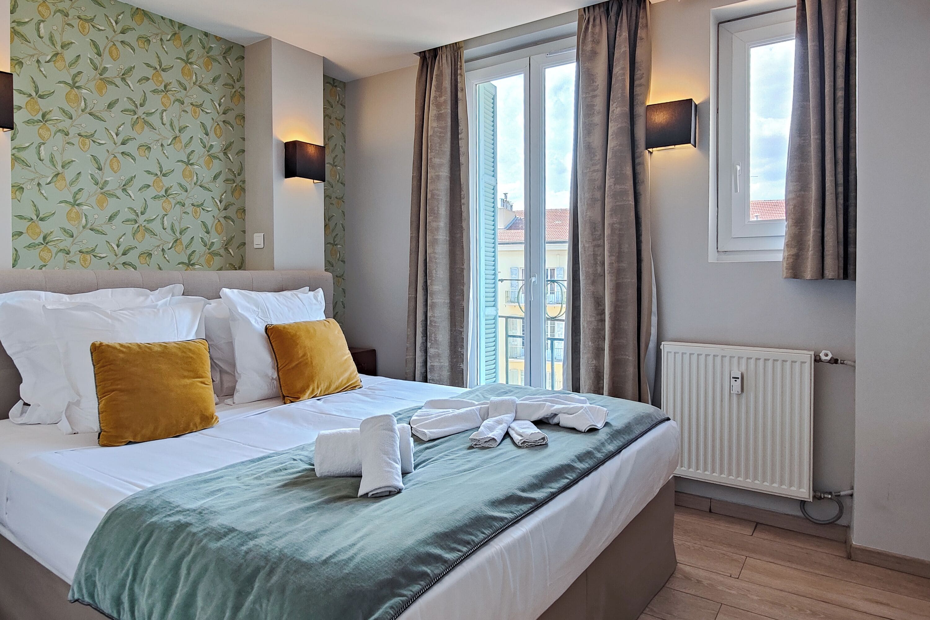 A hotel room with a bed, white linens, a green throw blanket, and a view of buildings through a window with curtains.
