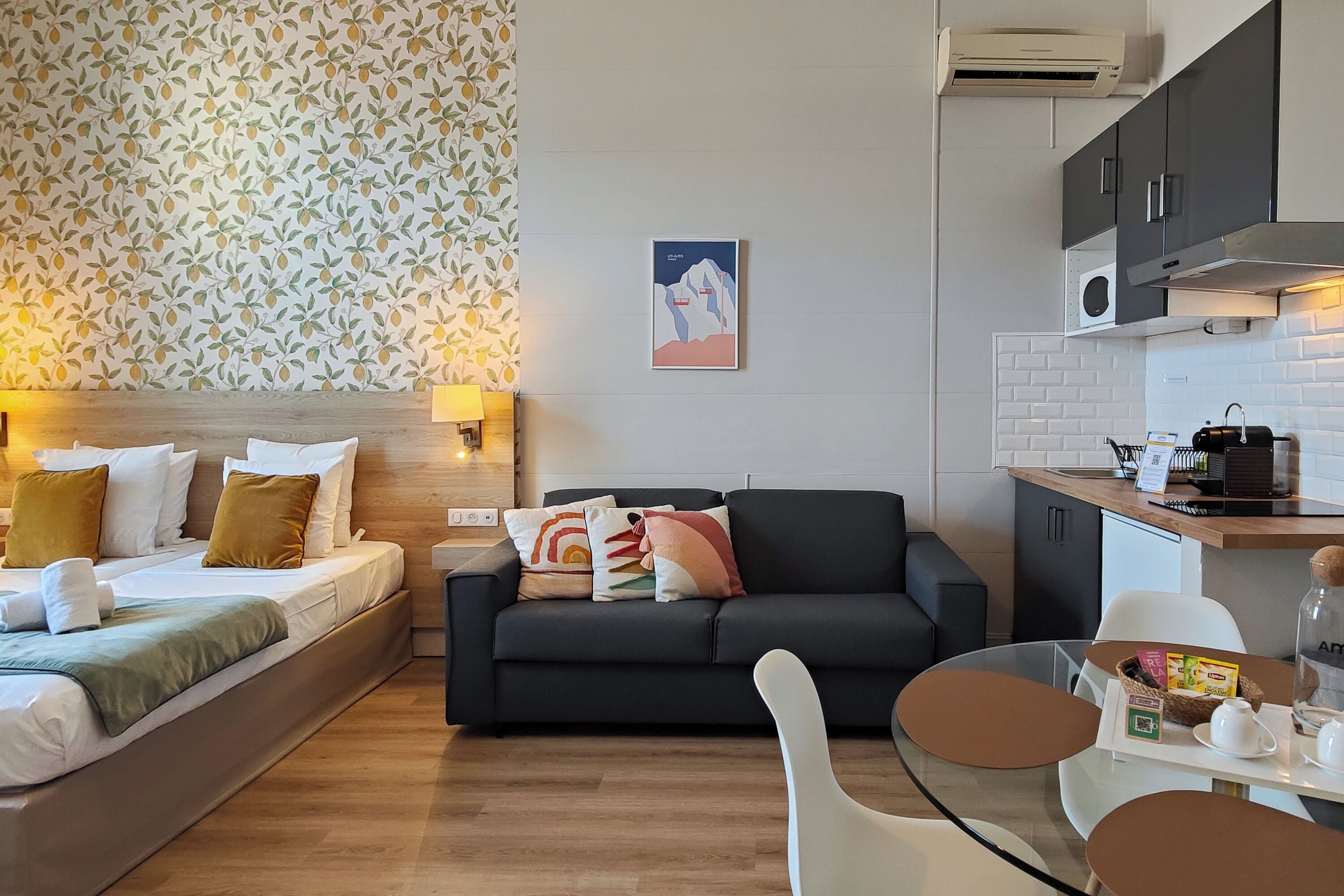 A modern hotel room with a bed, a sofa, a dining table, and a kitchenette.
