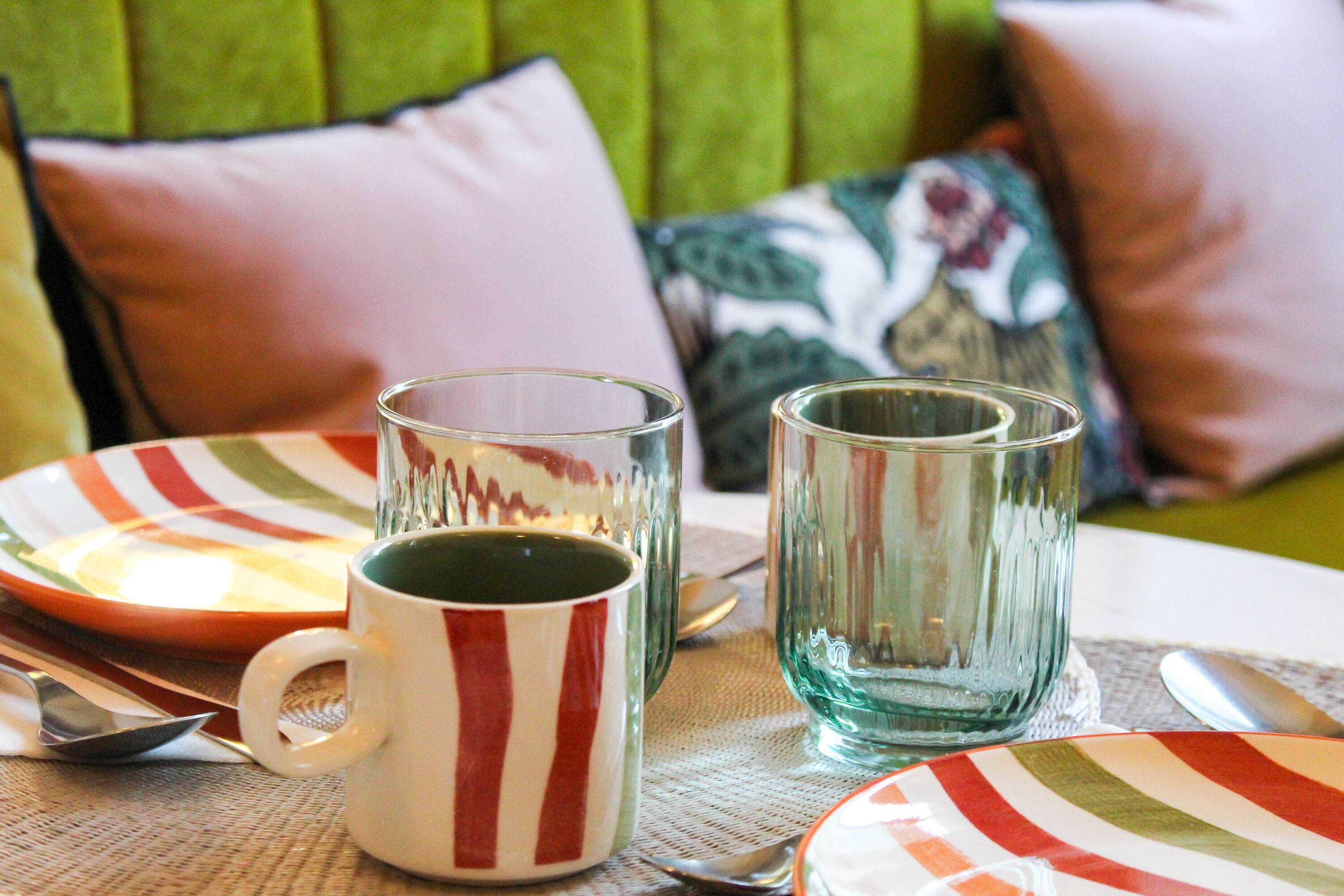Una mesa dispuesta con platos a rayas, una taza a rayas y vasos transparentes, con almohadones al fondo.