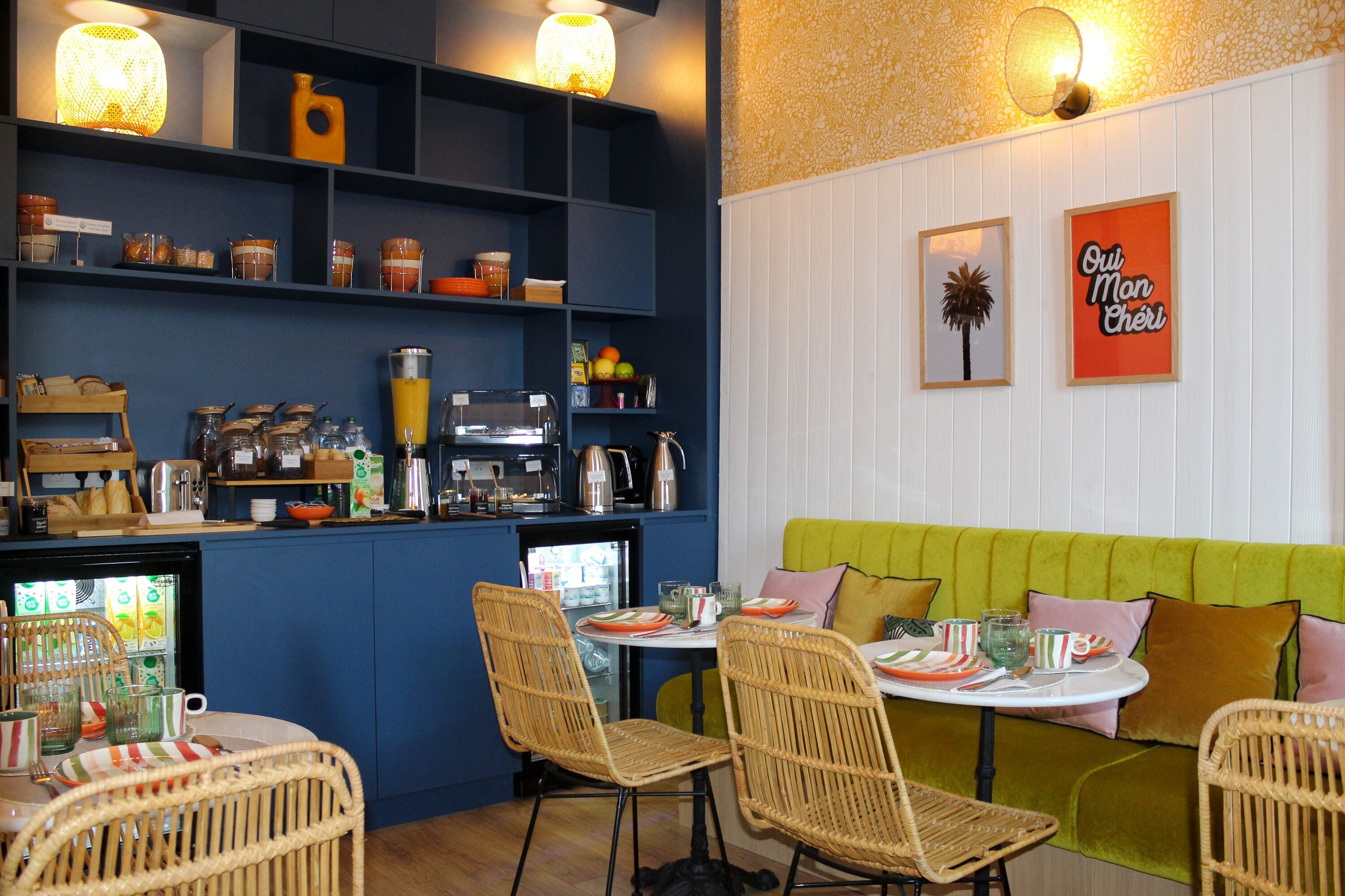 Un restaurant accueillant avec des chaises en osier, un coin salon vert et un espace cuisine doté d’étagères et d’appareils électroménagers.