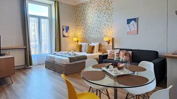 A modern hotel room with a bed, a sofa, a dining table with chairs, and a view of the outdoors through a window.