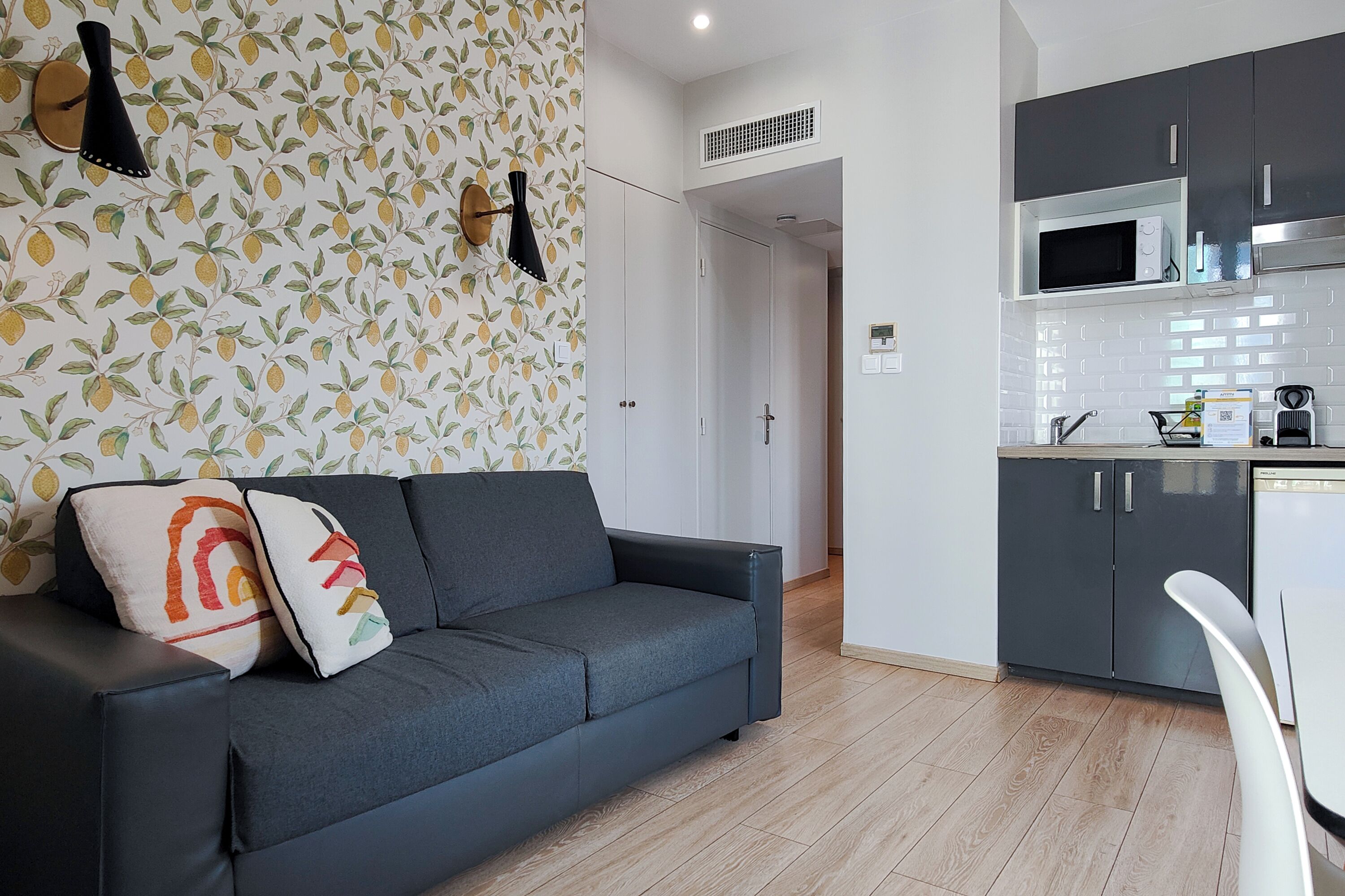 Un salon moderne avec un canapé gris foncé, un fauteuil blanc et un coin cuisine doté de meubles sombres et d’un four à micro-ondes.