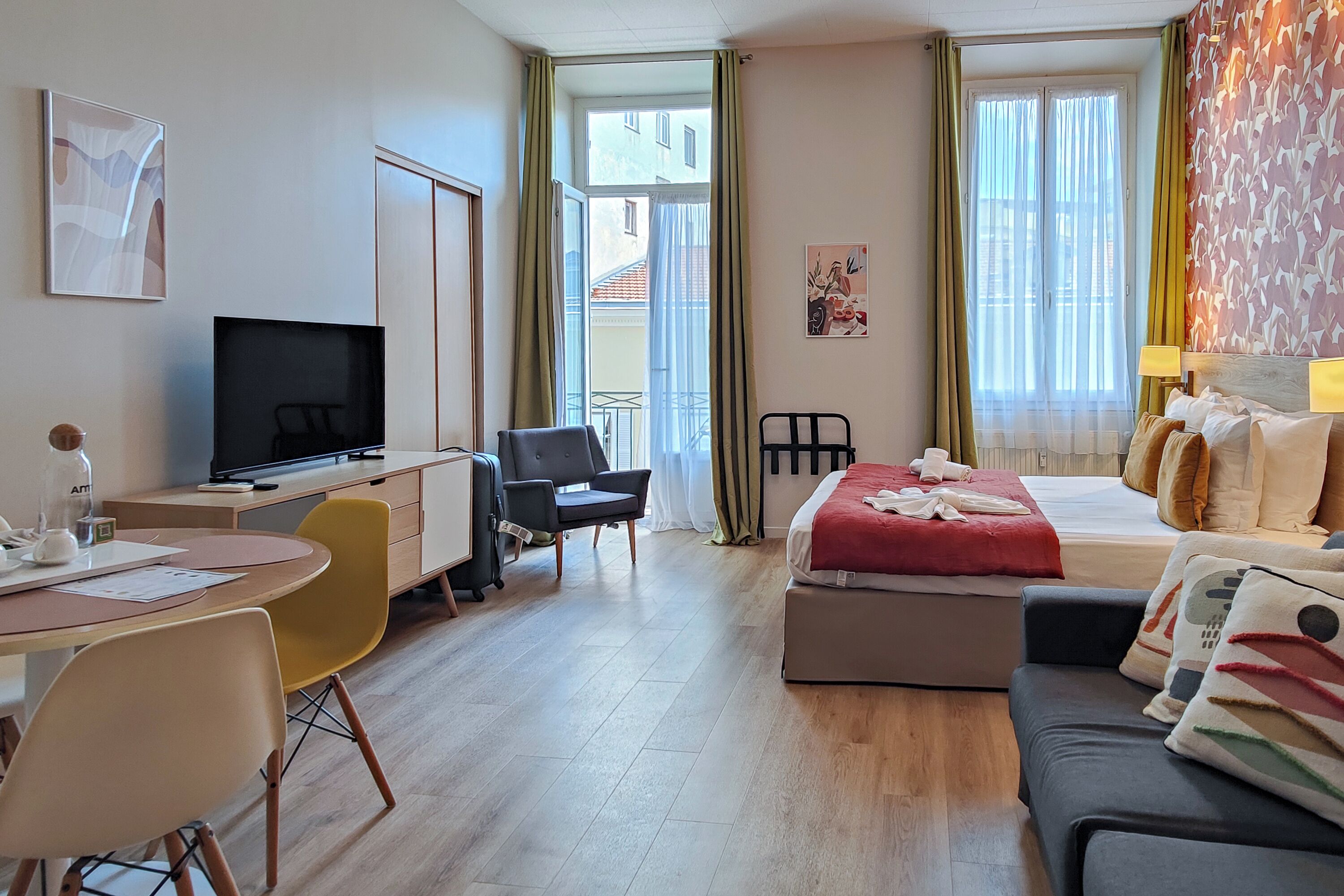 A hotel room with a bed, a desk with a TV, a chair, a sofa, and a balcony with curtains.
