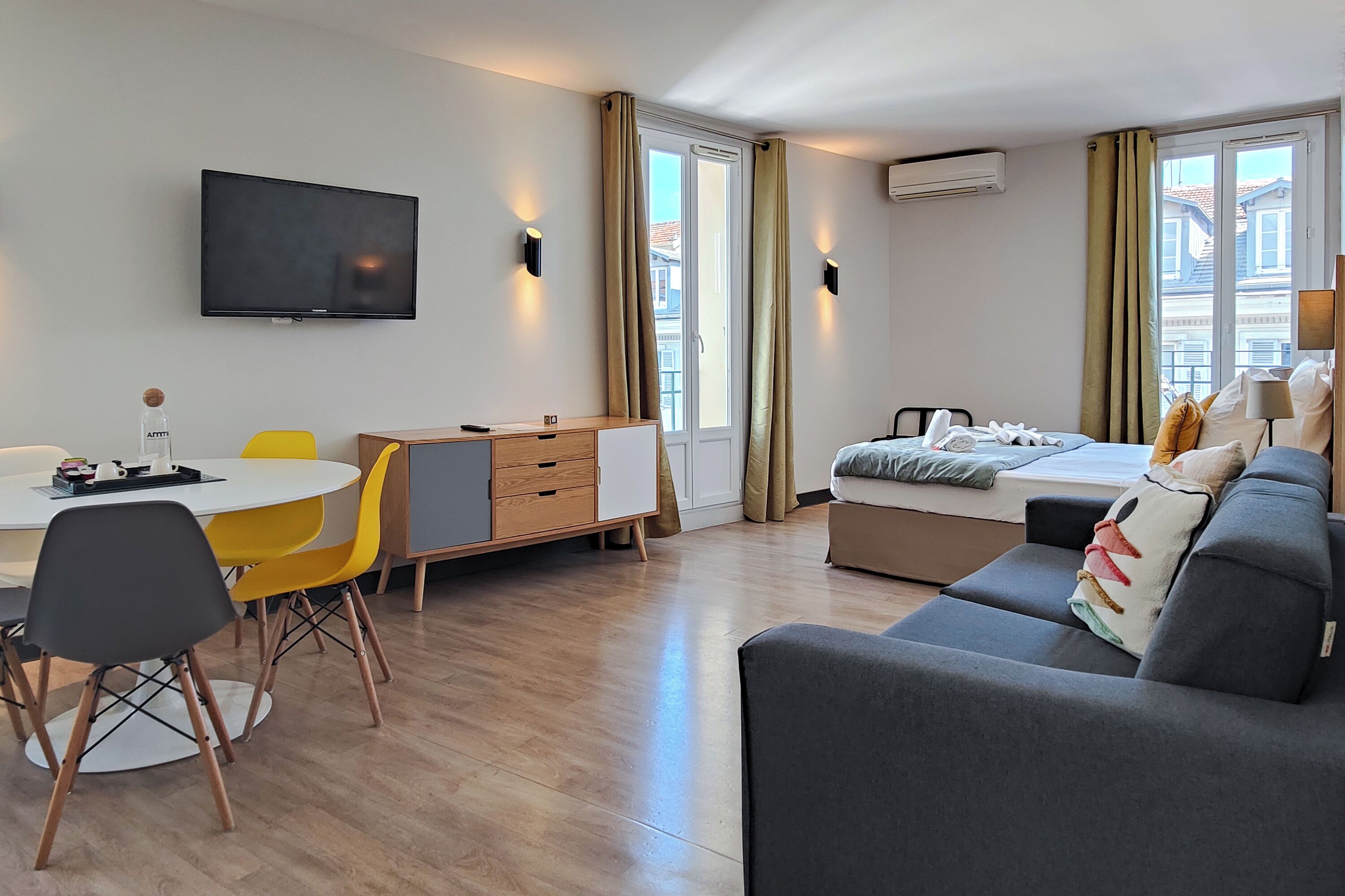 A modern hotel room with a flat-screen TV, a dining area with a round table and chairs, a sofa, a bed with pillows, and large windows with curtains.