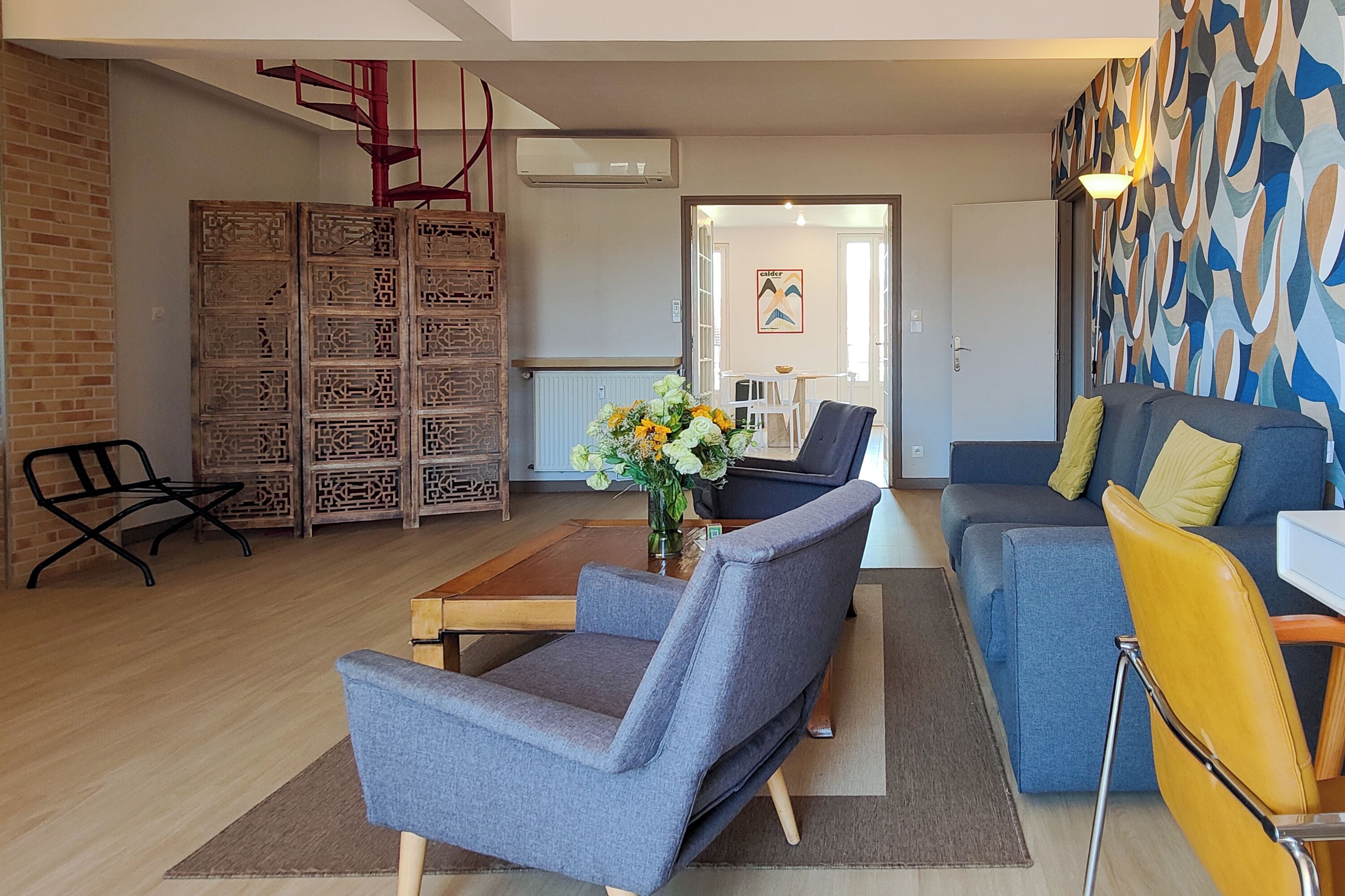 A modern living room with a grey sofa, a blue sofa, a wooden coffee table, and a black armchair. There is a brick wall on the left and a patterned wall on the right.
