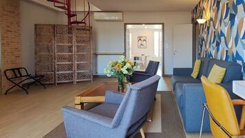 A modern living room with a grey sofa, a blue sofa, a wooden coffee table, and a black armchair. There is a brick wall on the left and a patterned wall on the right.