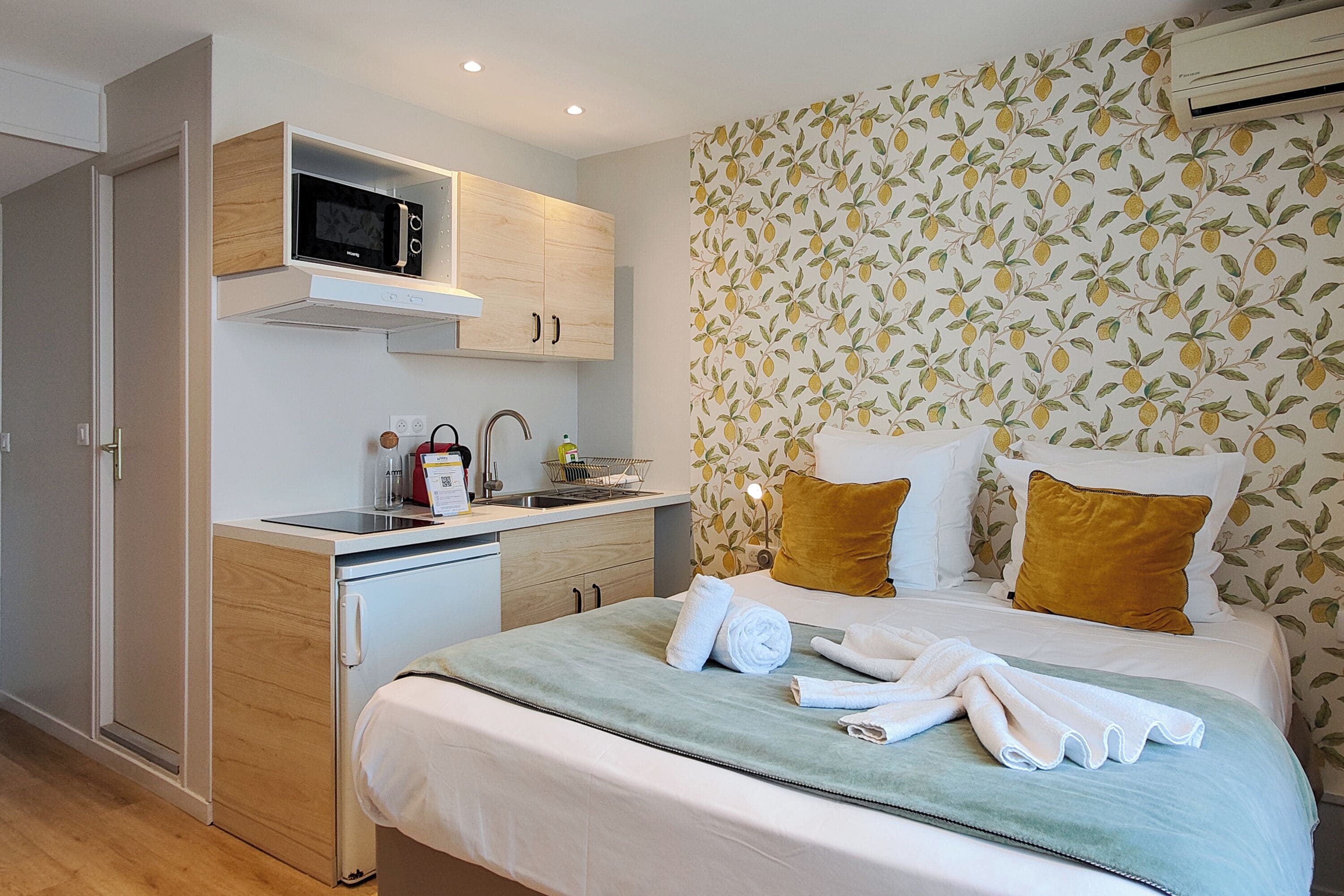 A compact hotel room with a kitchenette, a bed with two pillows, and a wall with a leaf pattern.
