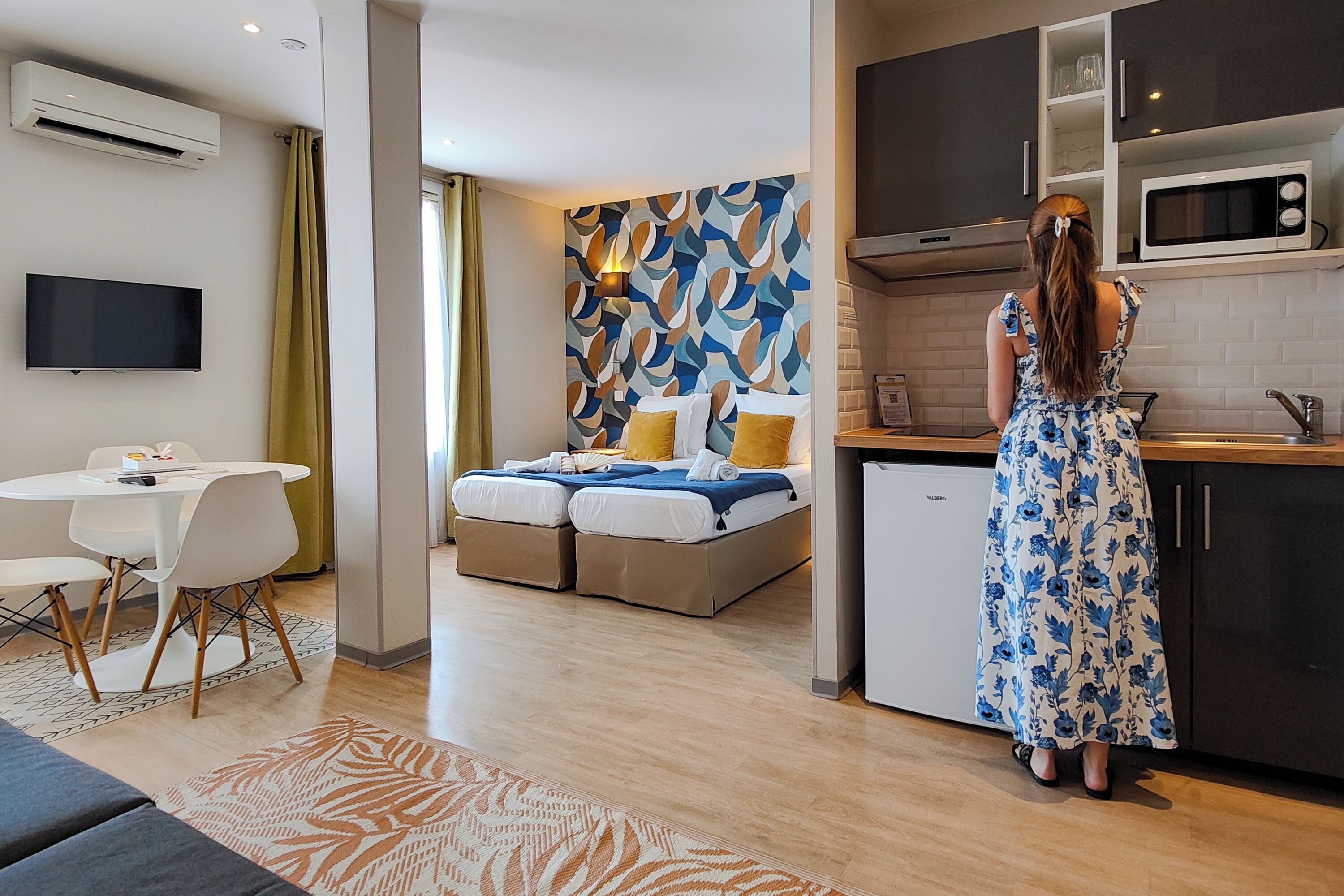 A modern hotel room with a bed, a dining area, and a kitchenette.