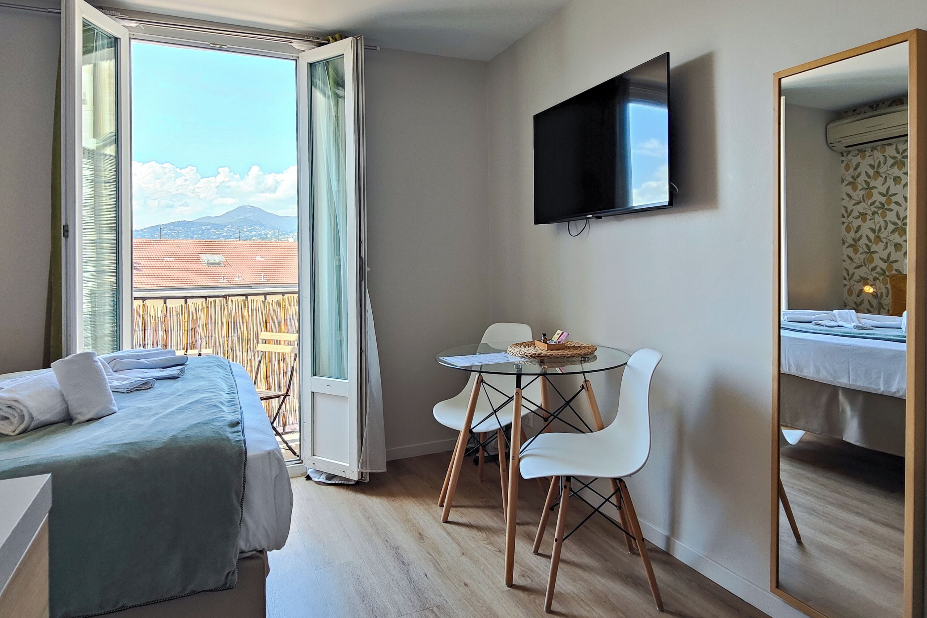 A hotel room with a bed, a TV, a dining table with chairs, and a balcony with a view of mountains.