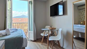 A hotel room with a bed, a TV, a dining table with chairs, and a balcony with a view of mountains.