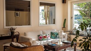 A cozy living area with a white sofa, a wooden coffee table, and a wicker chair. There are two large windows with curtains, a potted plant, and a framed picture on the wall. - Hotel Senaria (Anacapri)