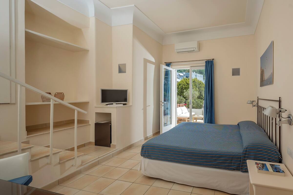 A bedroom with a bed, a TV, a desk, and a balcony with a view of trees.