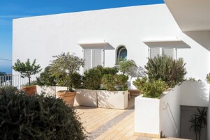 Un edificio blanco con dos ventanas y un pequeño arco, rodeado de plantas en macetas y una terraza de madera.