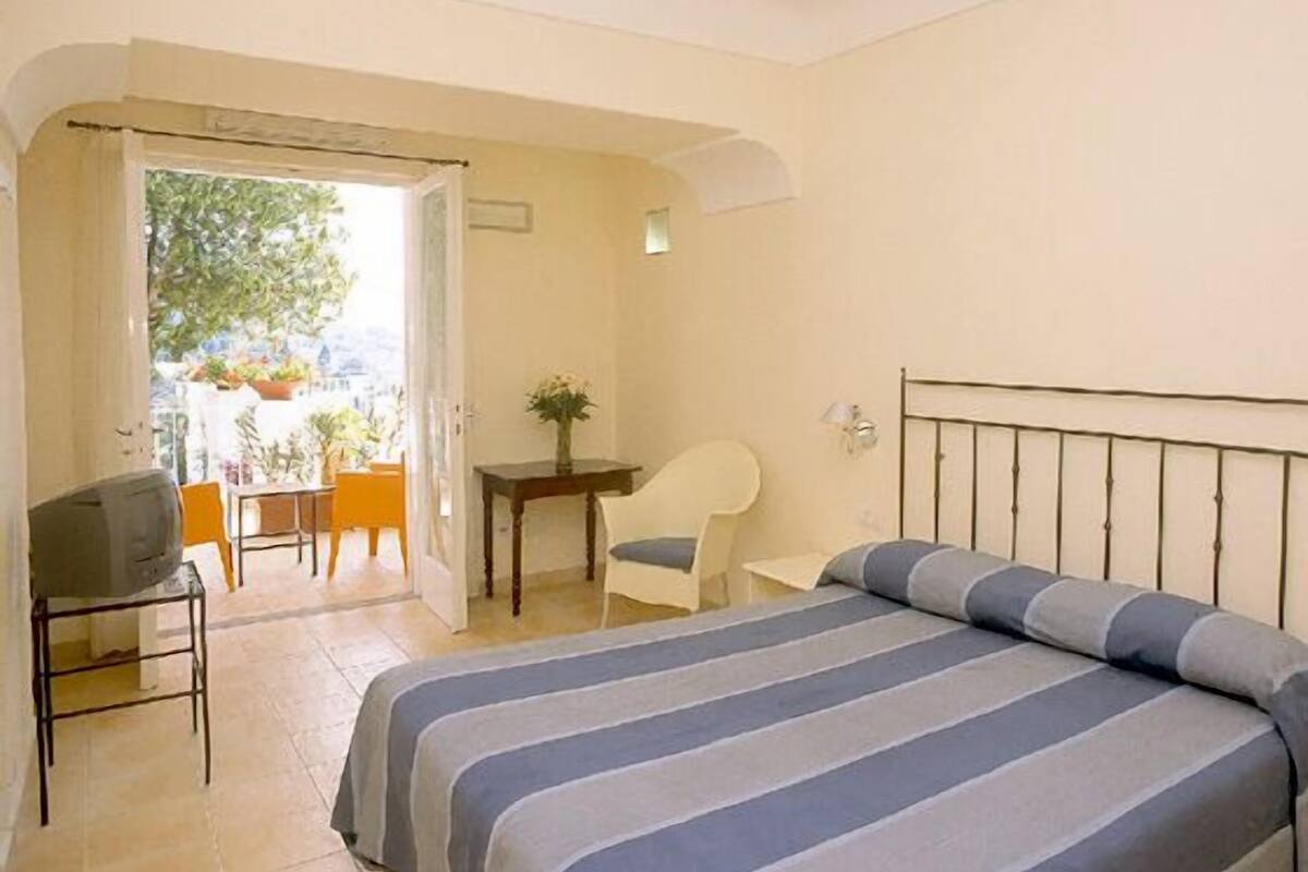 A bedroom with a bed, a desk, a chair, and a view of the outside.