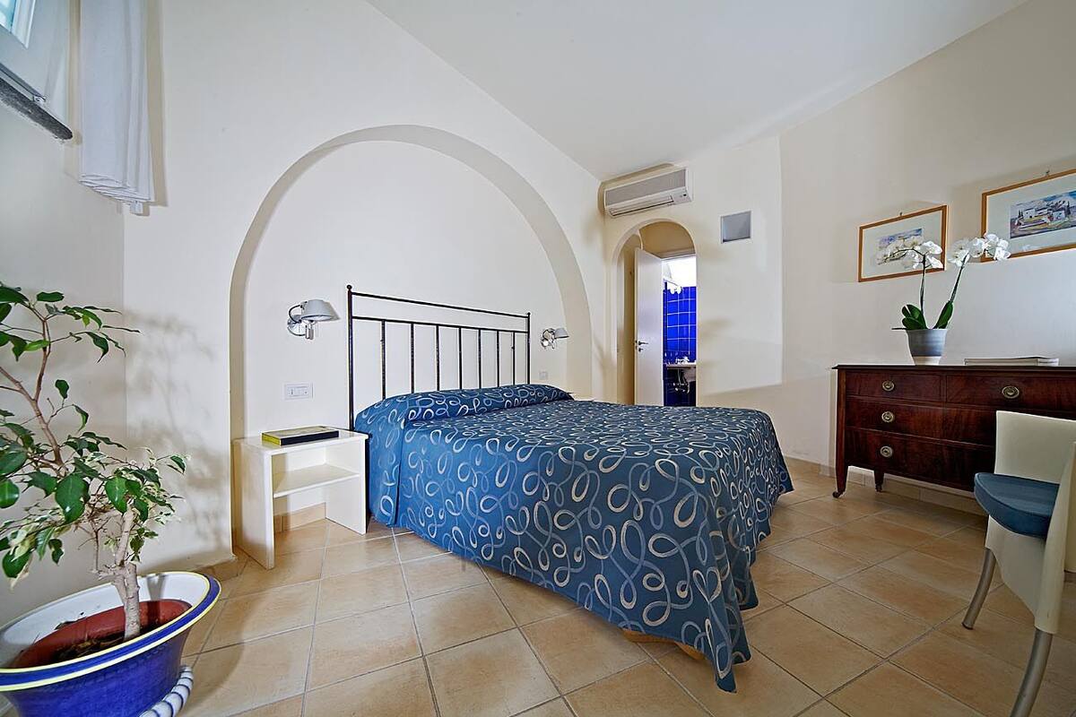 A bedroom with a bed, a desk, a chair, a potted plant, and a bathroom visible through an archway.