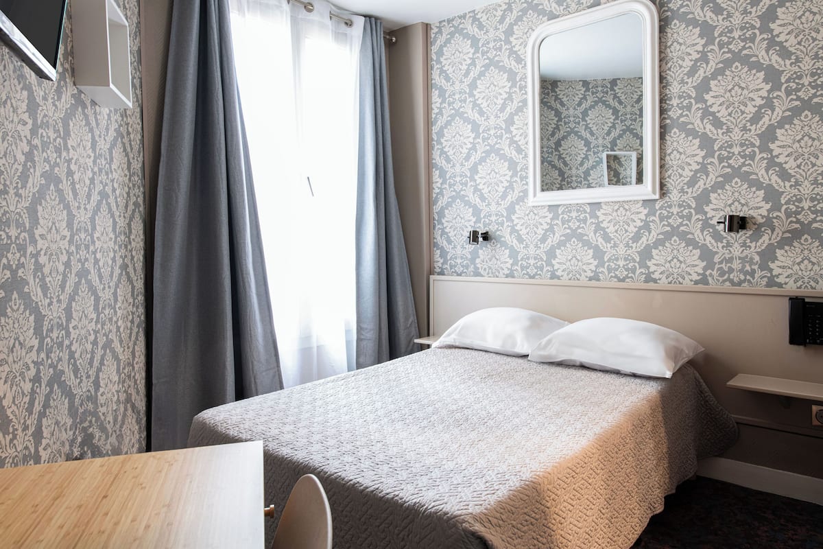 A hotel room with a bed, a bedside table, a chair, a mirror, and a window with curtains.