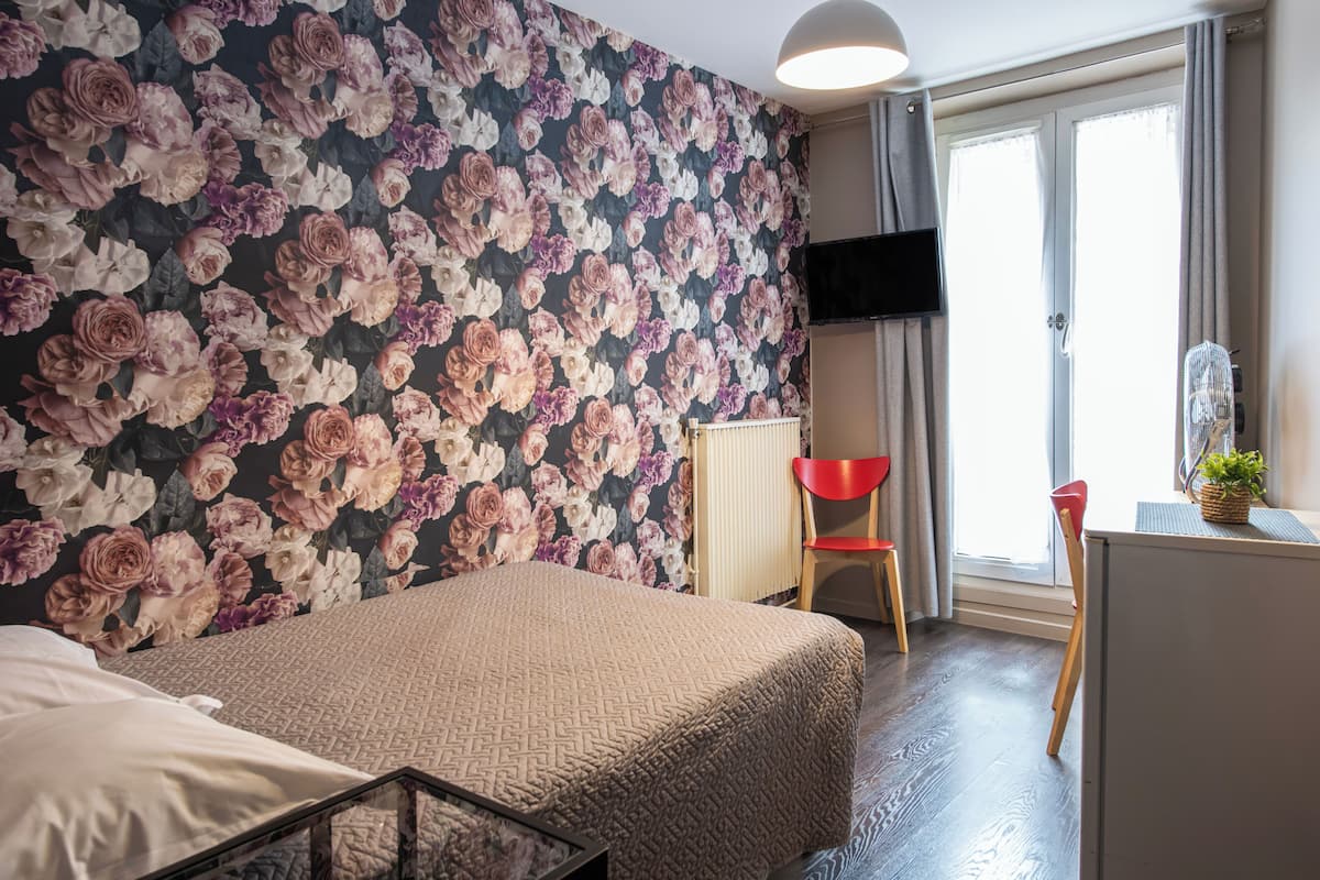 A bedroom with a floral wallpaper, a bed with a patterned bedspread, a red chair, a desk with a plant, and a TV on the wall.
