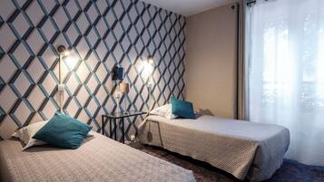 Habitación de hotel con dos camas, un papel tapiz decorativo, una mesita con lámpara y una ventana con cortinas translúcidas.