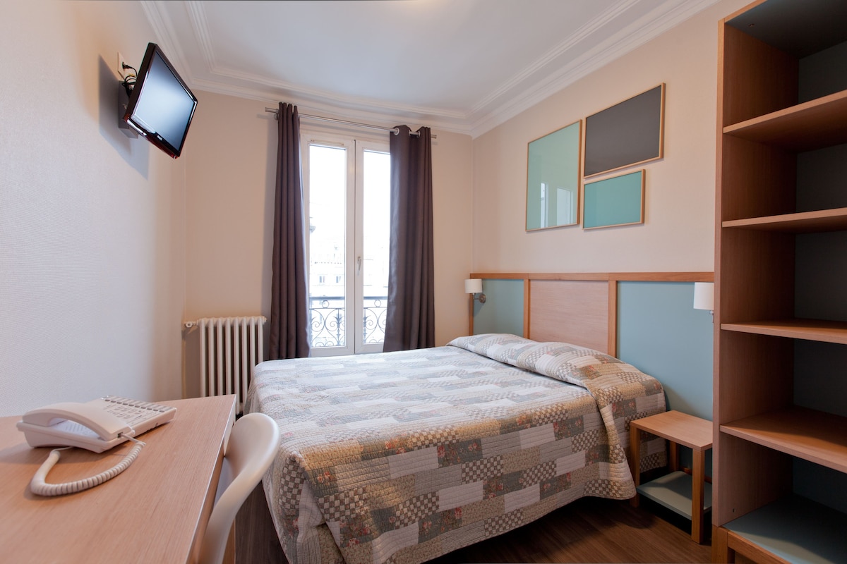 Une chambre d’hôtel comprenant un lit, un bureau avec un téléphone, un téléviseur fixé au mur et une fenêtre avec des rideaux.