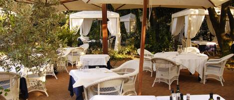 An outdoor patio with white wicker furniture, tables covered with white tablecloths, and umbrellas providing shade.