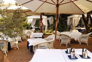 An outdoor patio with white wicker furniture, tables covered with white tablecloths, and umbrellas providing shade.