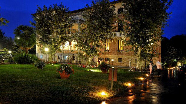 A historic building with a courtyard, illuminated by street lamps, surrounded by lush greenery and potted plants. - Park Hotel Villa Ariston (Camaiore)