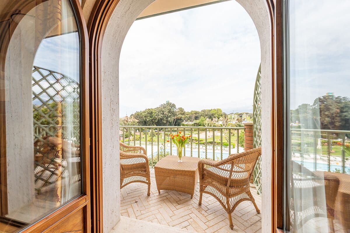 Ein Balkon mit Korbmöbeln, einem Tisch mit Blumen sowie Blick auf Garten und Pool.