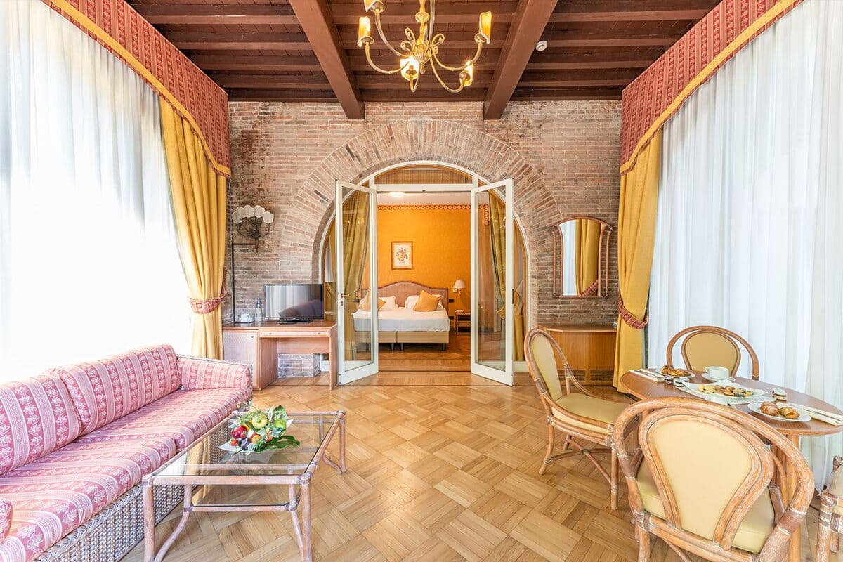 Ein Hotelzimmer mit Backsteinwand, Holzboden, einer Couch, einem Esstisch und einem Bett mit Blick durch eine offene Tür.