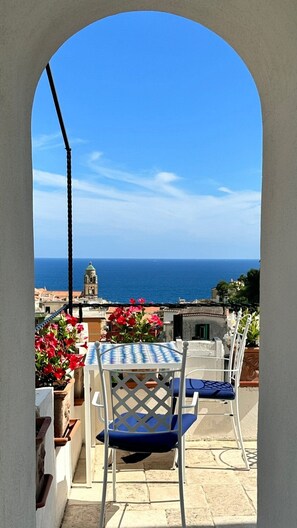 Una terraza con una mesa y sillas, vista al mar y un edificio con un campanario en el fondo.