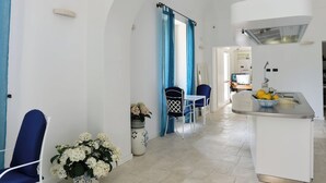 Una cocina moderna con encimera blanca, un lavamanos y una ventana. Hay una zona de comedor con una mesa y sillas, y se ve una silla azul.