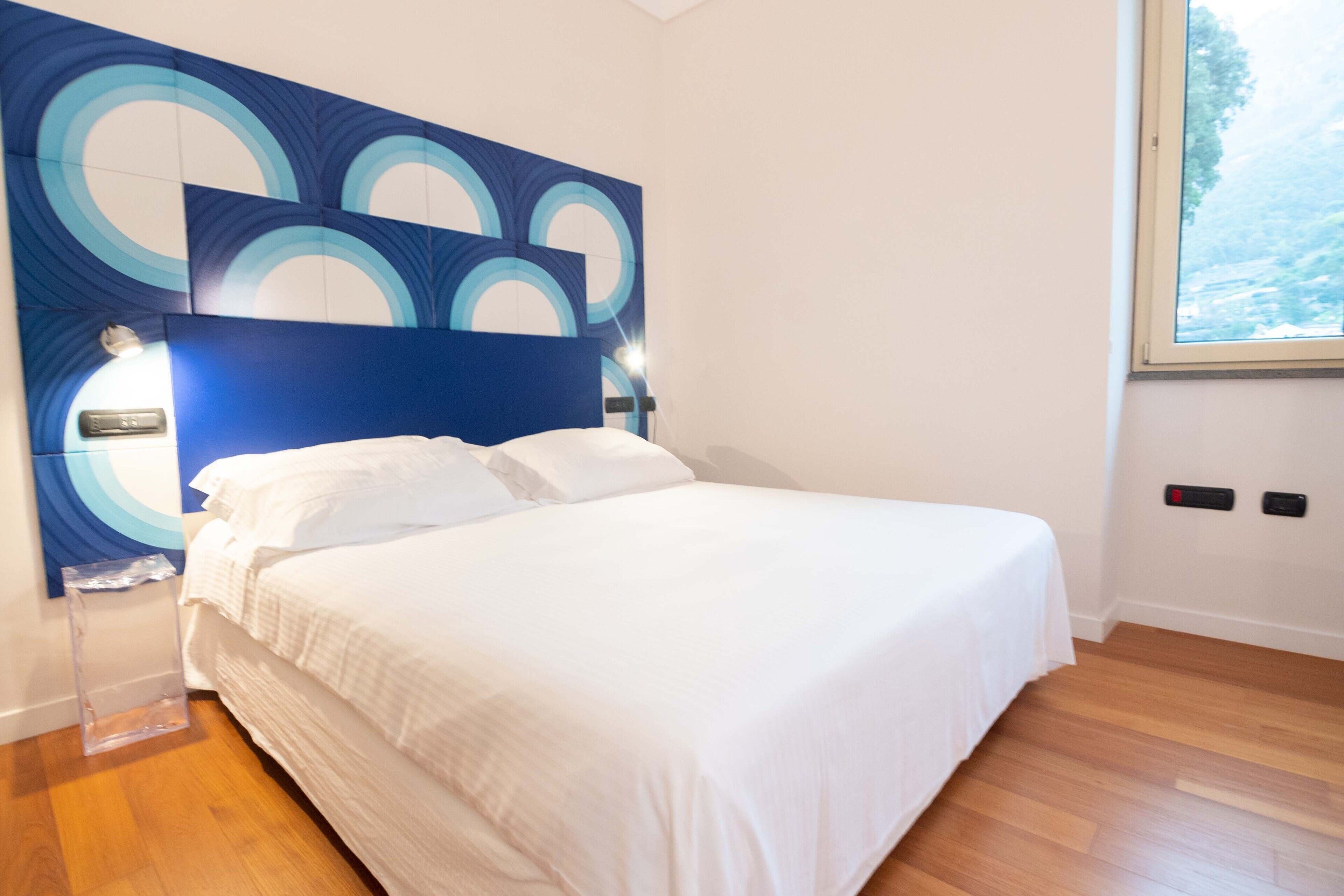 Un lit bien fait, avec du linge de lit blanc, une tête de lit bleue ornée de motifs circulaires et une fenêtre donnant sur un paysage verdoyant.