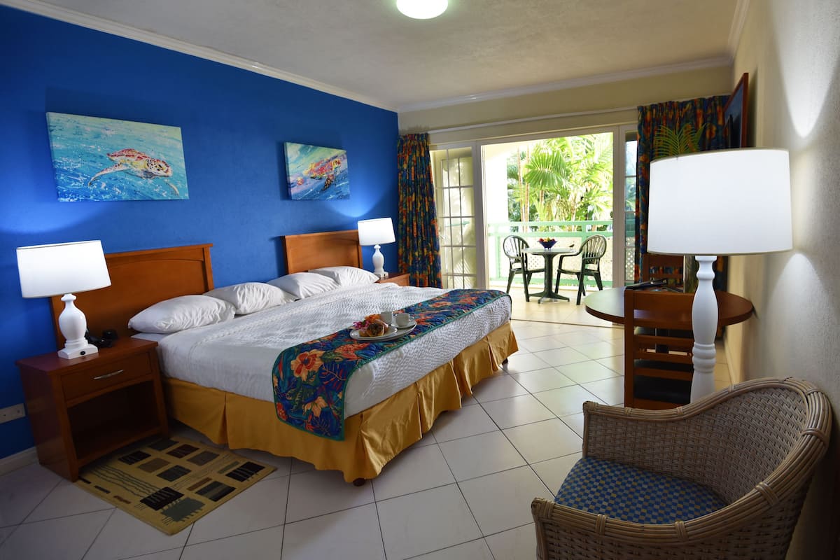 A hotel room with a bed, two bedside tables with lamps, a wicker chair, a small balcony with a table and chairs, and a view of greenery outside.