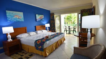 A hotel room with a bed, two bedside tables with lamps, a wicker chair, a small balcony with a table and chairs, and a view of greenery outside.