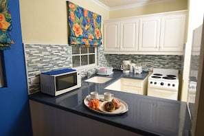 A kitchen with white cabinets, a microwave, a stove, and a window with a colorful curtain. - Blue Orchids Beach Hotel (Worthing)