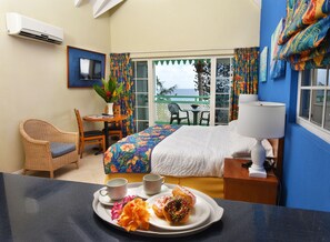 A hotel room with a bed, a desk with a chair, a table with a chair, a window with curtains, and a tray with donuts and flowers. - Blue Orchids Beach Hotel (Worthing)