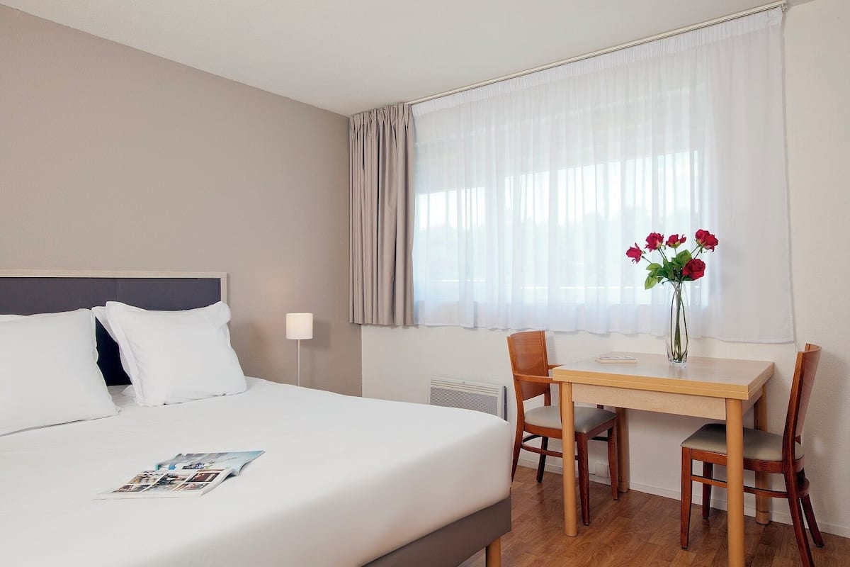 Habitación de hotel con cama, escritorio y silla. Hay una ventana con cortinas y un florero con flores en el escritorio.