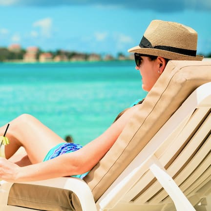 A person relaxing on a beach chair with a drink, overlooking a clear blue sea and a distant shoreline.