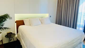 Una cama bien hecha con sábanas blancas, una mesita de noche, una planta en maceta y vistas al exterior a través de una ventana con cortinas.
