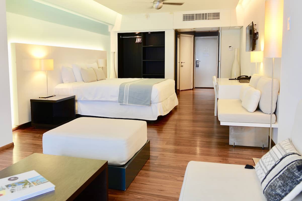A modern hotel room with a bed, a sofa, a coffee table, and a desk. There is a closet with hanging clothes and a door leading to another room.
