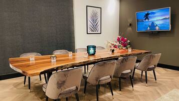 Un salón de conferencias con una larga mesa de madera, seis sillas, un jarrón con flores, una obra de arte con una hoja enmarcada y un televisor en la pared.