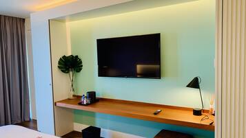 A modern hotel room with a large flat-screen TV mounted on the wall, a wooden desk with a remote control, a small black stool, and a potted plant.