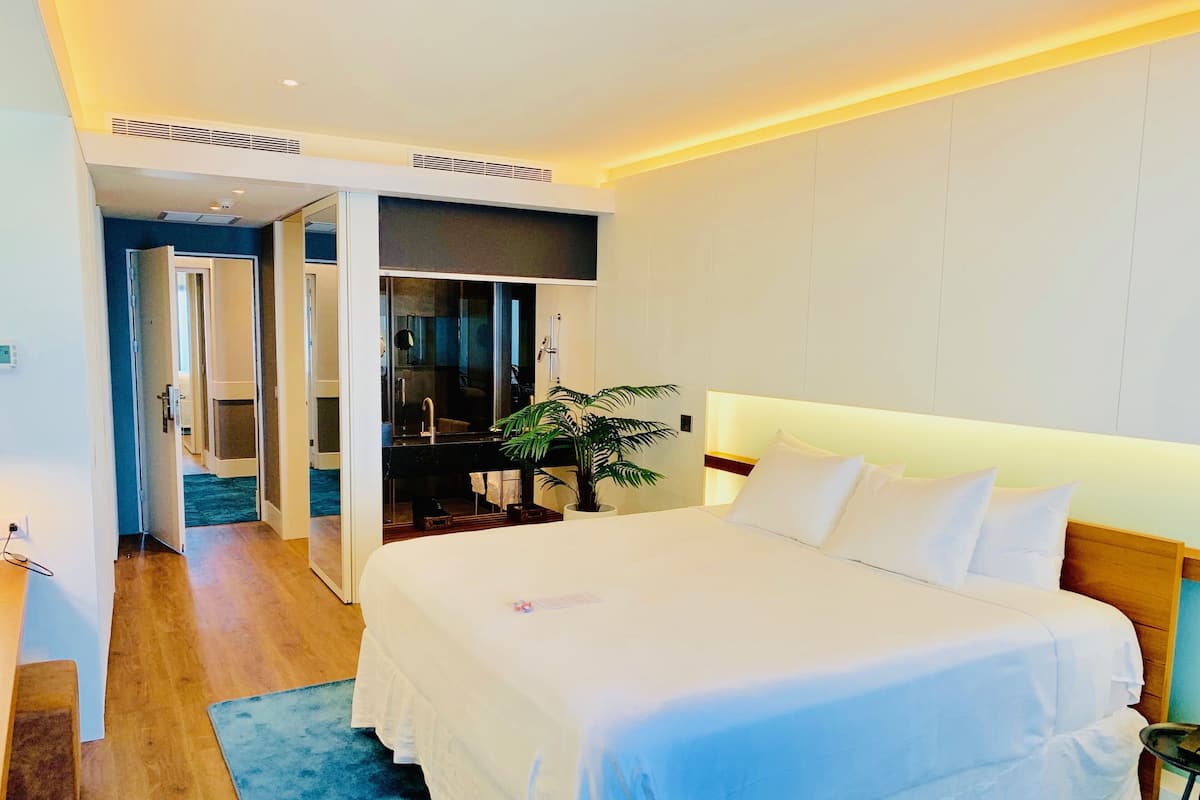 A hotel room with a large bed, a bedside table, a potted plant, and a view of a bathroom and hallway.